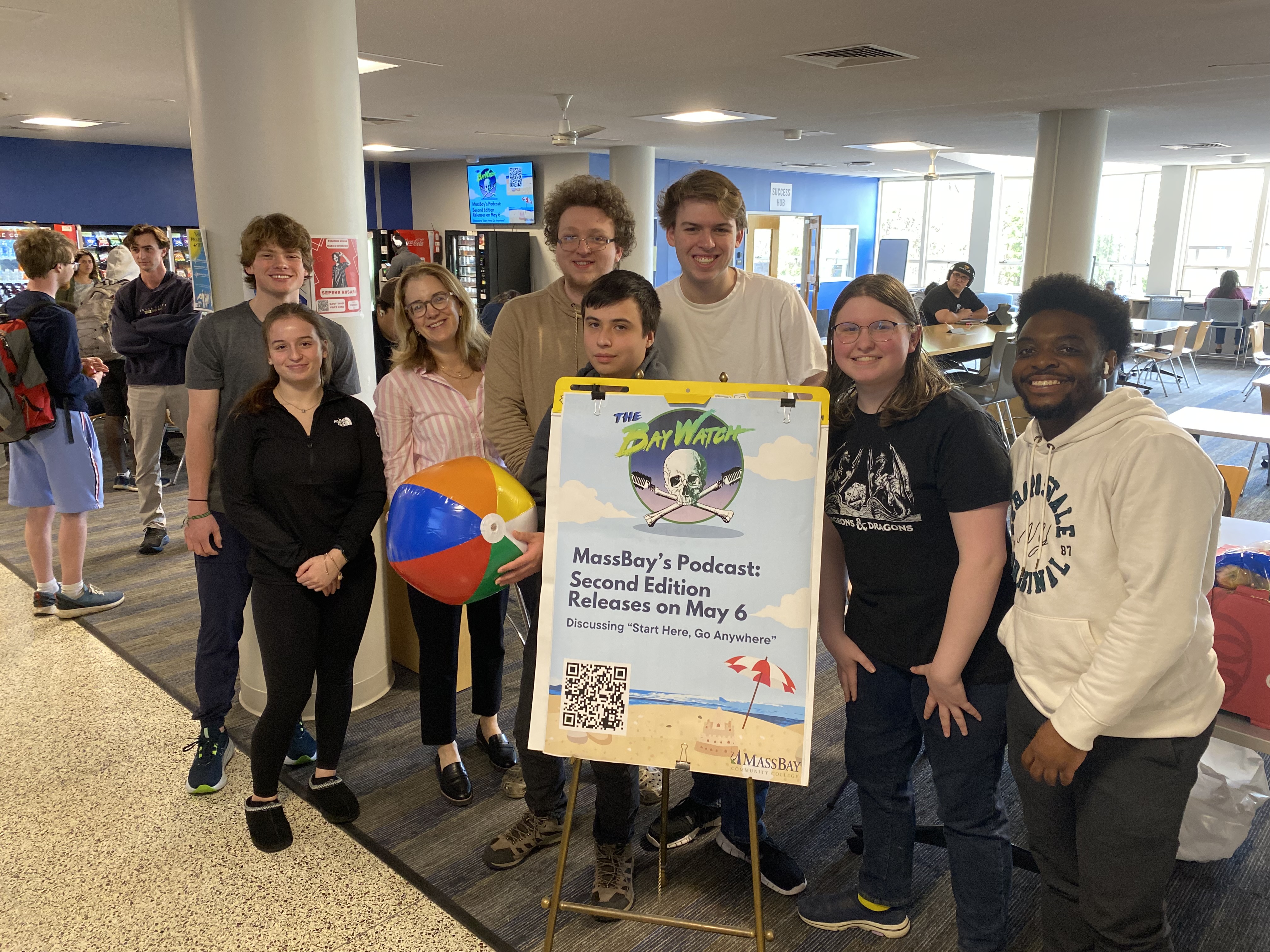 Seven student interns and professor standing around sign on stand advertising spring 2024 podcast. 